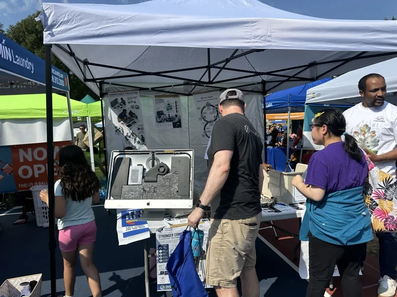 Community members inspecting a disassembled iMac and electronics display at an outreach booth